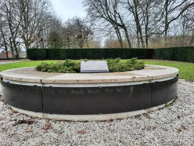 The image shows a memorial in the middle of a park with a plaque on it, surrounded by plants,...