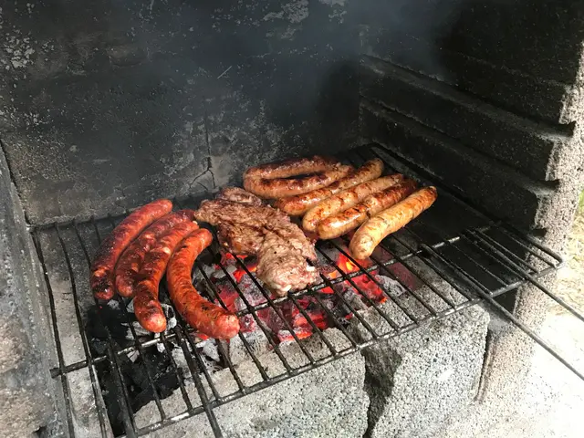 The image shows a barbecue grill with sausages and meat cooking on it, surrounded by coal and fire....