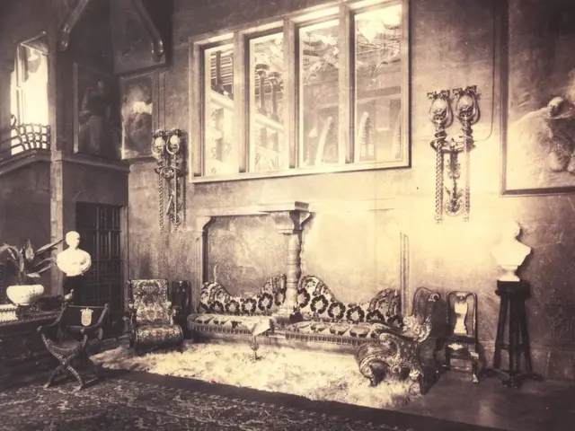 The image shows an old black and white photo of a living room with a studio couch, carpet, chairs,...