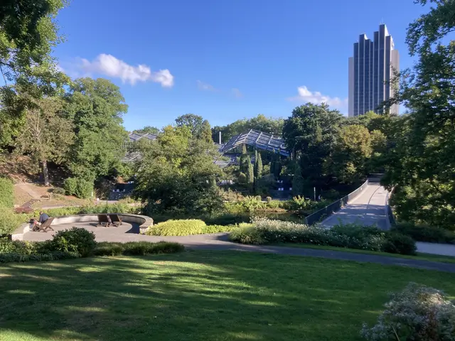 The image shows a park with lush green grass, trees, plants, and flowers, as well as benches and a...