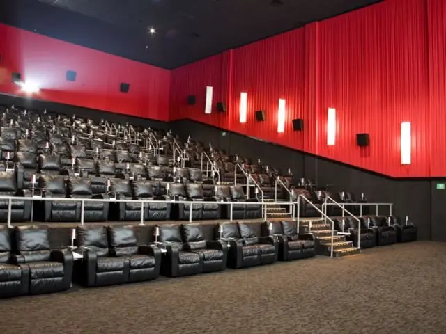 The image shows a large auditorium with rows of black leather chairs, a red curtain, steps with...